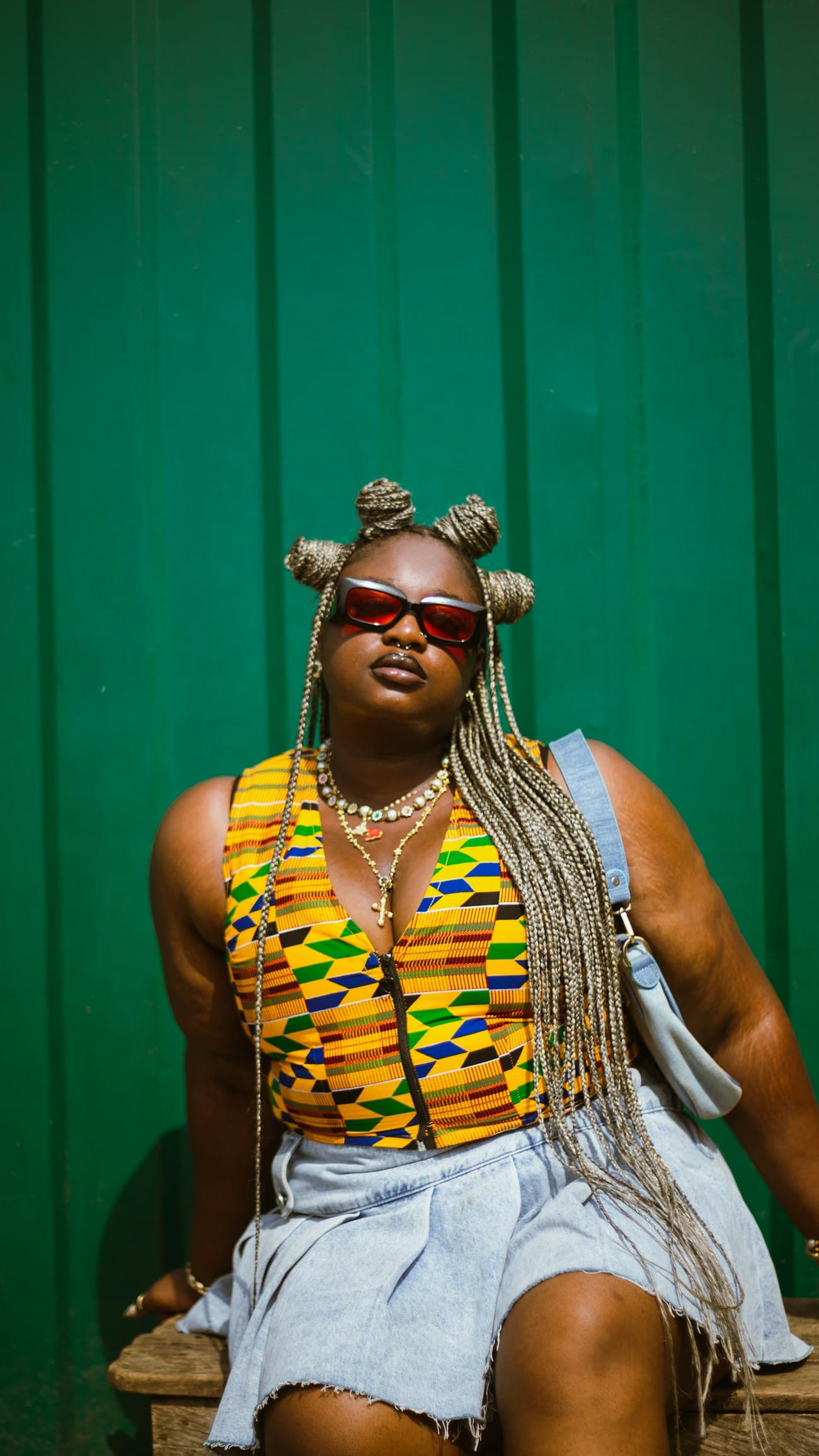 A woman in a kente top sits against a green wall